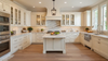 Bright luxury white kitchen with marble countertops, large island, farmhouse sink, glass-front cabinets, and hardwood floors